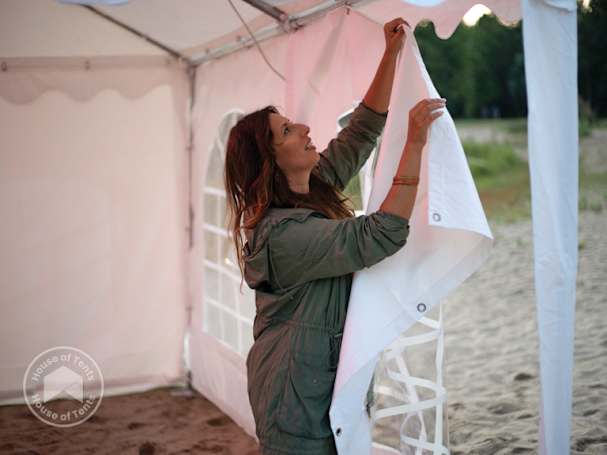 Woman attaching a white side panel to a 3x4 marquee on a sandy beach, demonstrating easy setup and weather protection.